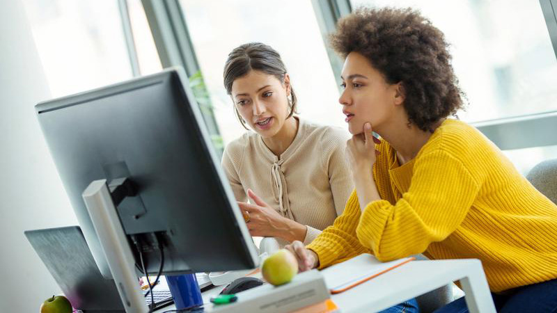 Two women look at a computer screen while sitting at a desk